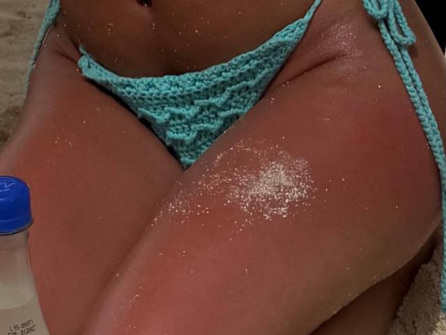 ChristieGarci's tan, sandy thighs in a teal, crocheted bikini bottom, with a blue-lidded bottle in the bottom left. Her skin is slightly shiny with sand.