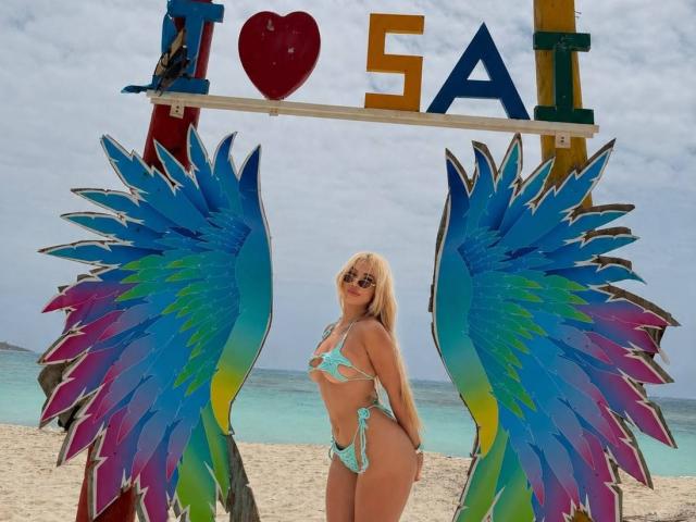 ChristieGarci with blue-green-feathered wings, wearing a teal bikini, standing on a sandy beach, her ass and tits visible, blonde hair, smiling, against a cloudy sky.