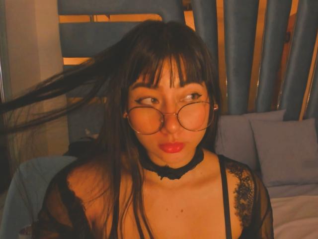 Genesiss, met lang zwart haar, een bril, rode lippenstift, een zwarte kanten top en een choker, frumt en houdt haar haar omhoog. Haar borsten zijn zichtbaar door het kant..