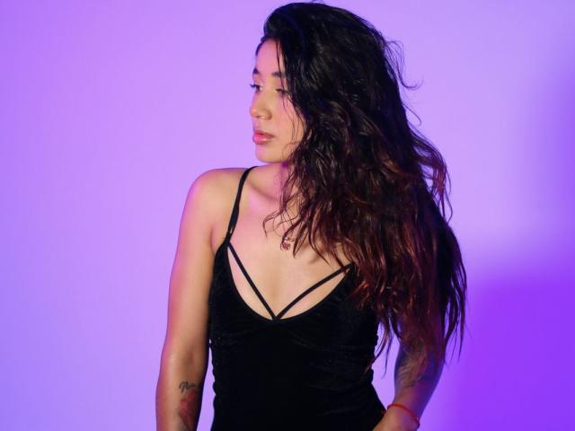 Erinsh with long, wavy dark hair, wearing a black strappy dress, showing tattoos on her arms, looking to the side with a slight smile, purple lighting.