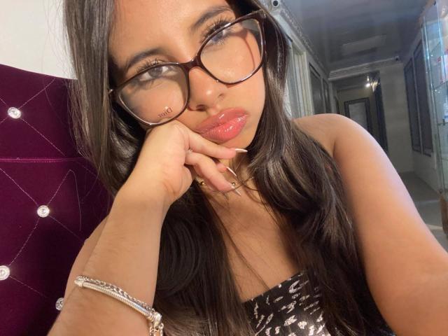 Celeste Morel, avec des lunettes, des lèvres roses, de longs cheveux noirs, tenant son visage, portant un haut noir et blanc sans bretelles, une bague à un doigt, un bracelet, un repose-tête violet avec des touffes..
