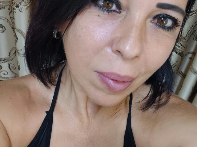 LauraSin's face, showing brown eyes with eyeliner, pink lips, and black hair, wearing a black halter top, with visible collarbone and earrings. Her skin is light and slightly shiny.