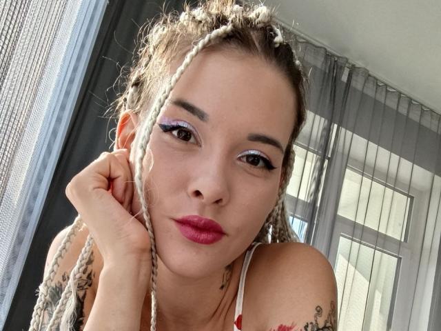 RachelNervis with braided blonde hair, purple eyeliner, pink lipstick, tattoos on her shoulder, and a hand touching her cheek, wearing a red and white top.