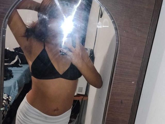 Mirror selfie of BonnieBodn with black bra, white pants, flash obscuring face, exposing toned midriff and small tits, brown skin, and a navel piercing.