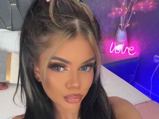 EvieBliss has blonde and black hair in a ponytail, full makeup with dark eyebrows, long eyelashes, and glossy pink lips. Neon "love" sign is in the background.