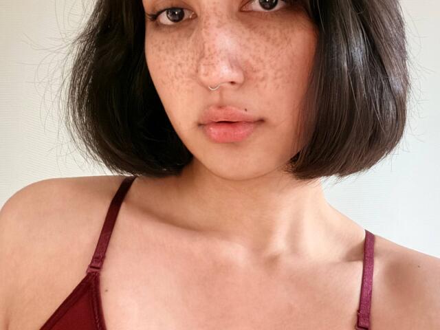 HeikeCarchi with freckled face, nose ring, and short black bob, wearing a red spaghetti strap top, showing her bare shoulders and collarbone. Her lips are slightly parted.