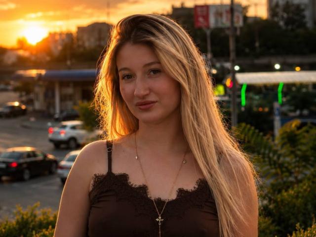 HollyTaillor with long blonde hair, wearing a brown lace top, gold necklace, and subtle makeup, standing in front of a sunset, smiling gently, with cars and greenery in background.