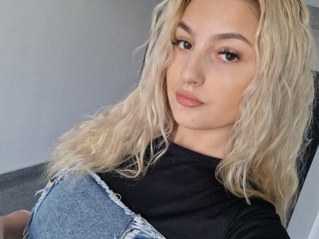 AngelofSinn with wavy blonde hair, brown eyes, and pink lips, wearing a black top and ripped blue jeans, sitting in a chair, looking at the camera.
