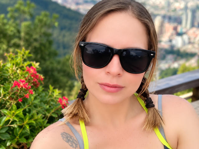 PamelaColin, avec des lunettes de soleil noires, des couettes blondes, un débardeur de sport gris néon, un petit tatouage sur l'épaule droite, des lèvres roses, une peau claire, du feuillage vert, des fleurs rouges, une rambarde en bois en arrière-plan..