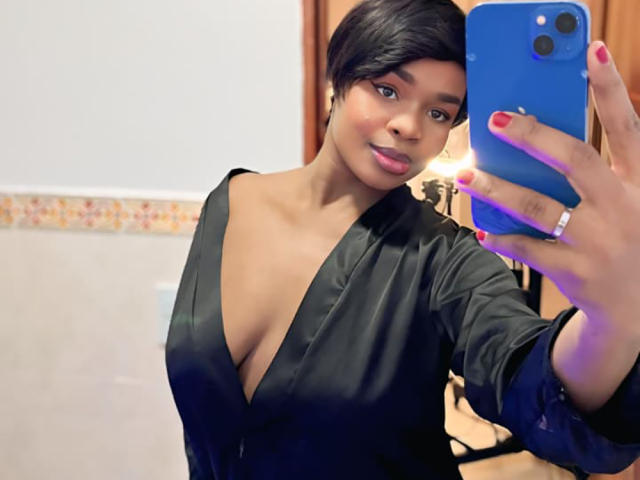VenusWilly with short black hair, wearing a deep black shirt, showing cleavage, holding a blue phone, red nails, ring, and smiling at the mirror.