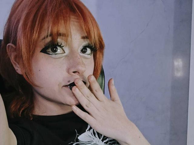ViolettClay, con i capelli rossi, l'eyeliner nero e un piercing al naso, che si tocca la bocca con un dito, mostrando un piccolo sorriso e un piercing al setto nasale..