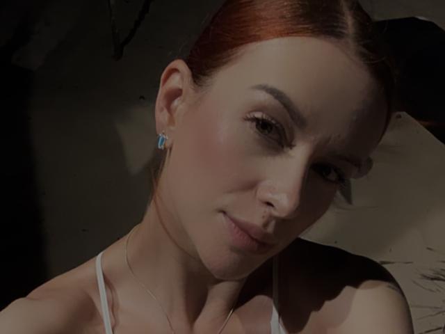 Leahred has red hair, light skin, and is wearing a white top. She has blue earrings, a slight smile, and is looking at the camera. Her tits are not visible.