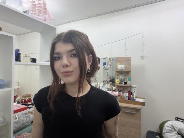 Julie Norberg mit braunen Haaren, einem schwarzen T-Shirt und Ohrringen, steht in einem Raum mit Make-up und Schönheitsprodukten, lächelt sanft und schaut in die Kamera..