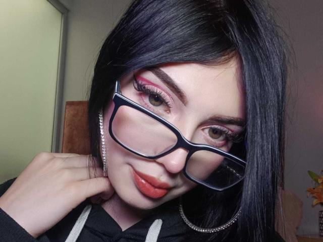 Isabella Rossi, con capelli neri, occhiali, ombretto rosa, rossetto rosso e un sorriso, tiene una mano al collo. Indossa una felpa nera con spalline bianche..