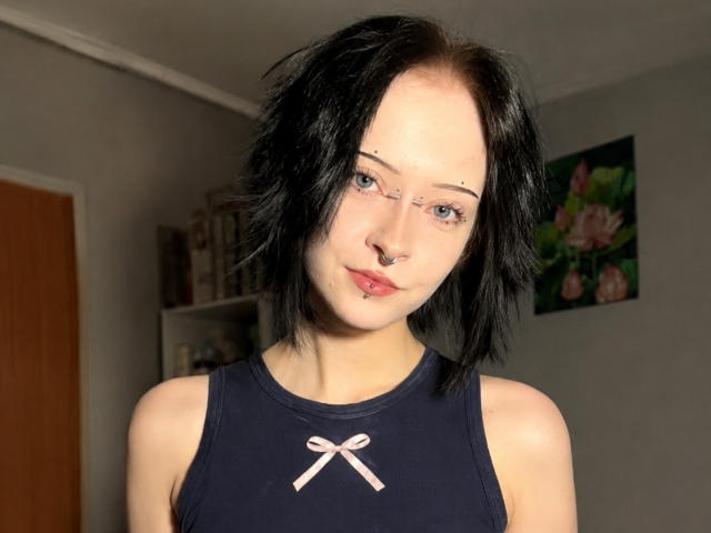 LianeBadenoch has black, messy hair, blue eyes, a nose ring, and a lip piercing. She wears a dark navy sleeveless top with a pink bow.