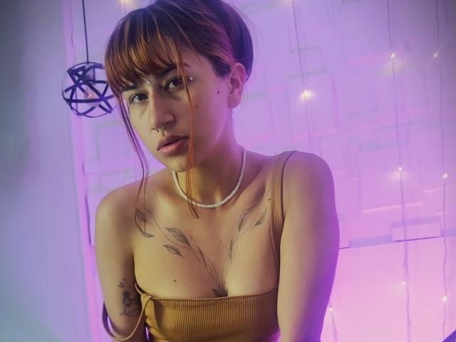 NaraStorn with brown hair, bangs, nose ring, and tattoos, wearing a brown ribbed top and pearl necklace, under purple and pink lights. Boobs, tattooed chest, looking at camera.