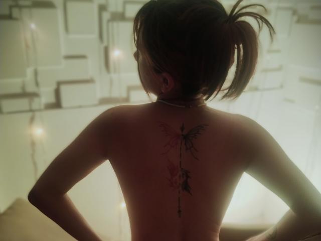 NaraStorn's back, topless, with a butterfly tattoo down her spine, hands on her hips, brown ponytail, subtle lighting, soft shadows, bare skin, slight blush, calm, confident.