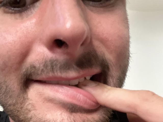 BlackMaskF's face showing a slightly open mouth with a finger gently touching his bottom lip. He has a light beard, brown eyebrows, and is slightly smiling.