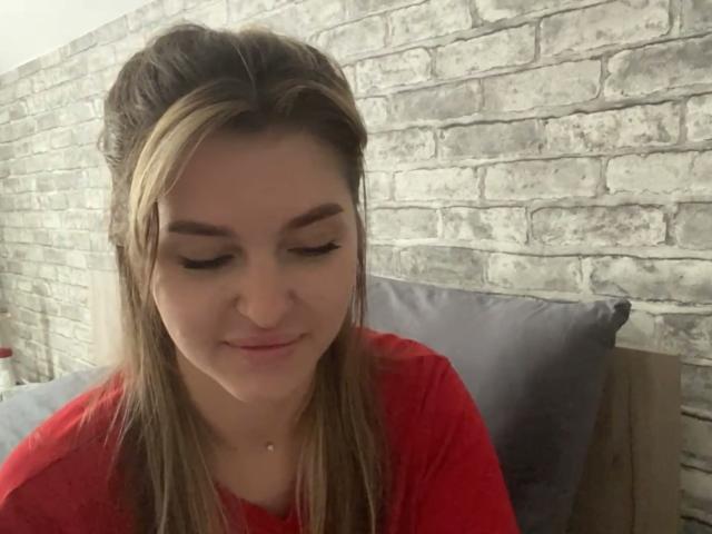 ElanaFalencki with long brown hair in a red shirt, looking down, slight smile, subtle makeup, small nose piercing, brick wall background, gray pillow behind her.