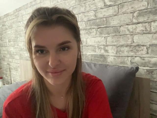 ElanaFalencki with long brown hair, wearing a red shirt, has small tits, and a cute face, smiling slightly, in a bedroom with a brick wall.