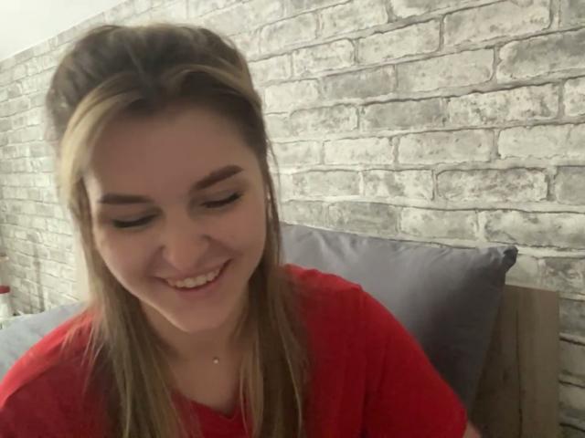 Elana Falencki a les cheveux blonds, elle sourit les yeux fermés, elle porte une chemise rouge et elle a un petit anneau au nez. Ses seins ne sont pas visibles. Elle est assise sur des coussins gris..