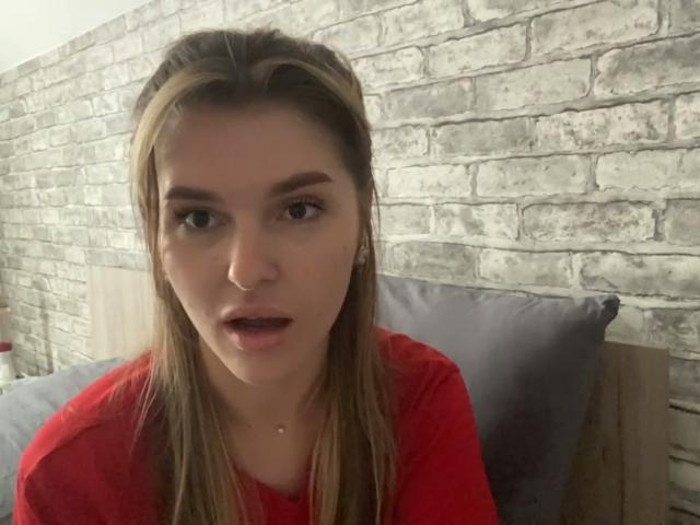 ElanaFalencki with straight brown hair, red shirt, and surprised expression, staring forward, mouth slightly open, in a dimly lit room.