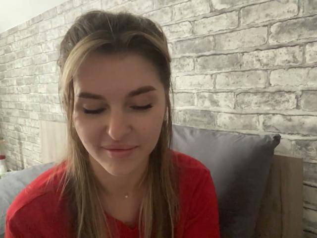 ElanaFalencki with long brown hair, wearing a red shirt, looking down with a slight smile, sitting on a gray pillow, brick wall in the background.
