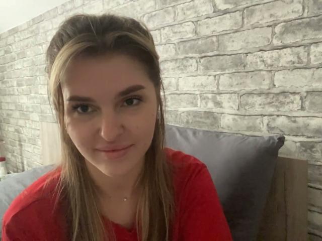 ElanaFalencki has light brown hair, red shirt, and a small nose piercing, smiling at the camera with gray pillows behind her. Her tits are not visible.