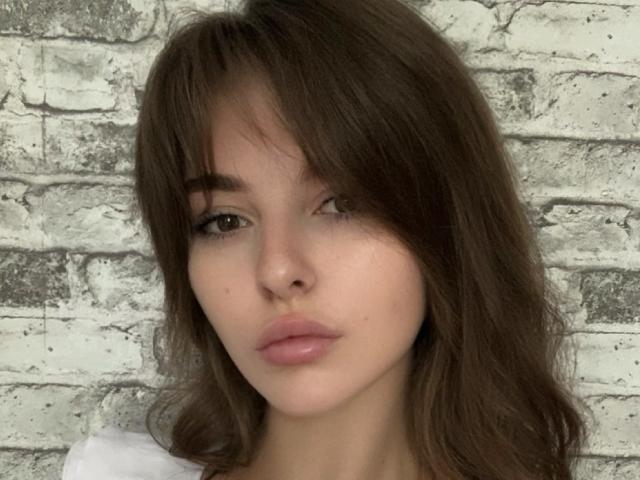 ElanaFalencki has wavy brown hair, full pink lips, and light brown eyes. She's looking slightly to the side with a neutral expression. Her white shirt is barely visible.