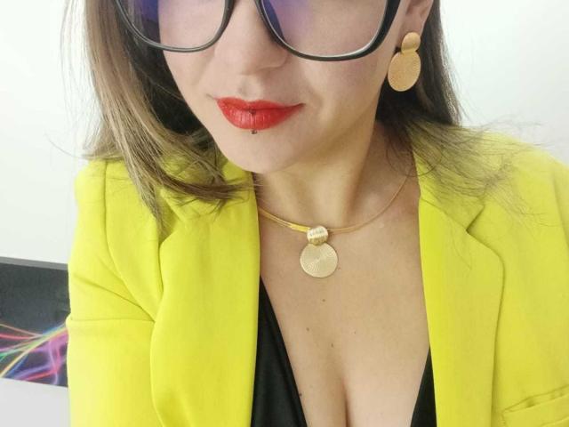 ArianaBlonde with bright red lipstick, black-rimmed glasses, yellow blazer, gold necklace, and black dress, showing her cleavage and small round earrings. Her boobs are slightly visible.