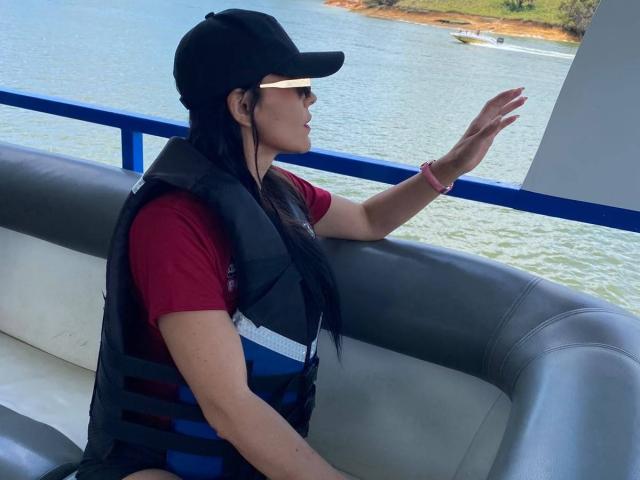 MindyLove with long black hair, wearing a black cap, red shirt, and black life vest, sits on a boat, waving with her right hand, looking outside.