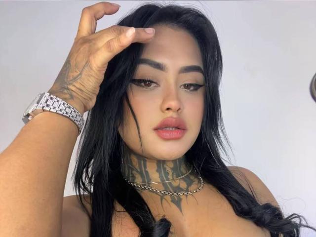LairaHearvy with black hair, tattoos on neck and hand, wearing a chain necklace and silver watch, looking down with hand in hair, showing off her tits and pouty lips.