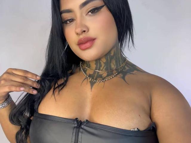 LairaHearvy with black hair, heavy eyeliner, and red lipstick, wearing a strapless leather top, showing tattoos on her neck, and a chain necklace, touching her hair.