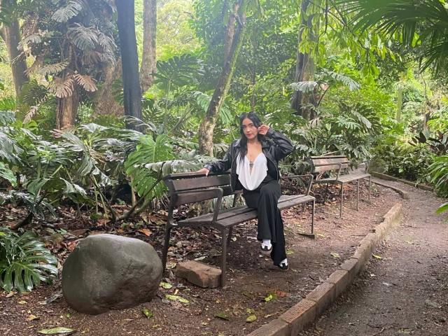 MindyLove with black hair, glasses, and a white blouse, sitting on a bench, wearing black pants, and white sneakers, in a lush green forest, next to a large rock.