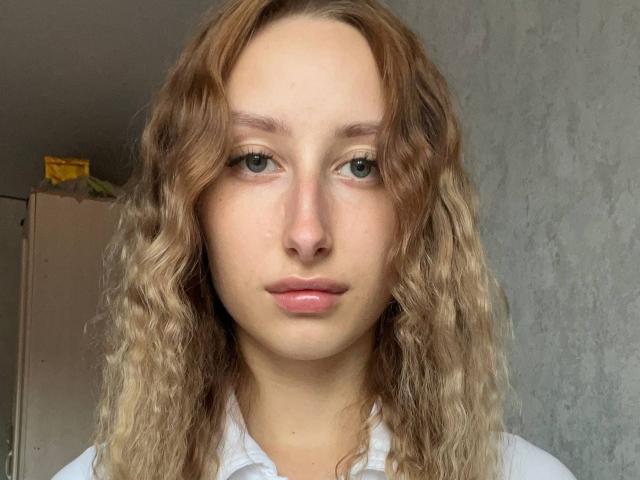 Isaura Latz ha i capelli biondi ondulati, gli occhi verdi e le labbra rosa, indossa una camicia bianca e ha un'espressione neutra in una stanza semplice con pareti grigie..