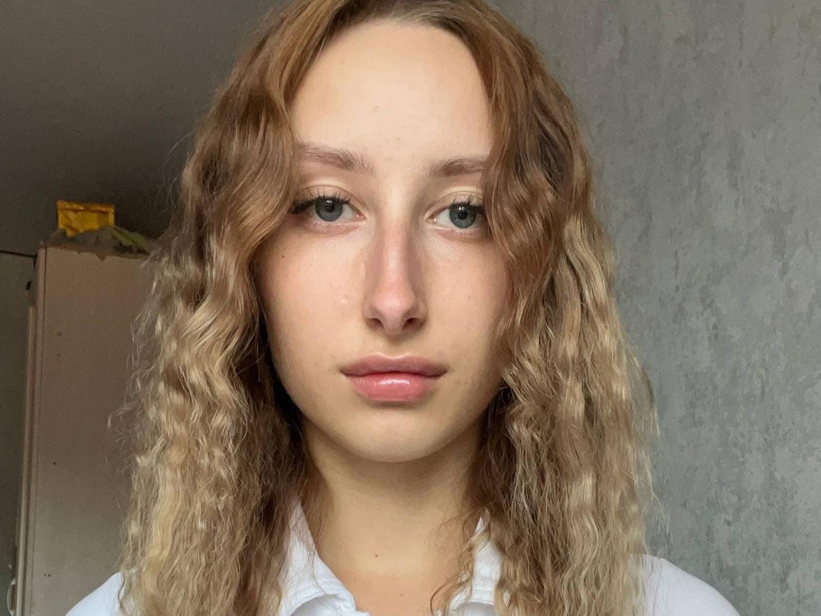 Isaura Latz ha i capelli biondi ondulati, gli occhi verdi e le labbra rosa, indossa una camicia bianca e ha un'espressione neutra in una stanza semplice con pareti grigie..