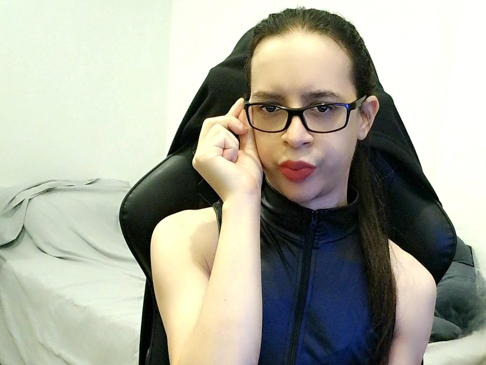 Noco1991 with glasses, red lipstick, black sleeveless zip-up, and black chair, touching glasses with right hand, long brown hair, pale skin, slight smile.