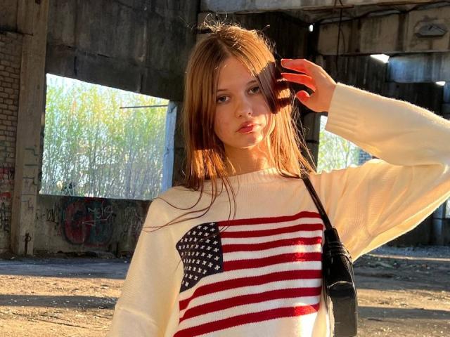 SuzieBrion with long brown hair, wearing a white sweater with a red and white American flag design, holding red headphones, standing under a sunlit urban structure.