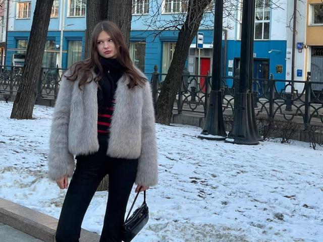 SuzieBrion with long brown hair, gray fur jacket, black turtleneck, red striped shirt, black pants, holding black purse, standing in snowy urban park, trees in background.