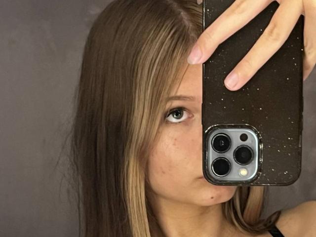 SuzieBrion with blonde hair, holding a phone in front of her face, showing one eye, and a small moleskin spot on her cheek. Her hand is on the phone.