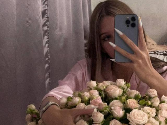SuzieBrion with long blonde hair, pink shirt, holding roses, taking selfie with blue phone, visible long white nails, holding flowers in front, partially covering face.