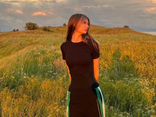 SuzieBrion stands in a field, wearing a black shirt and green-striped pants, with long brown hair, holding her arms, her tits subtly outlined, sunlight on her face.