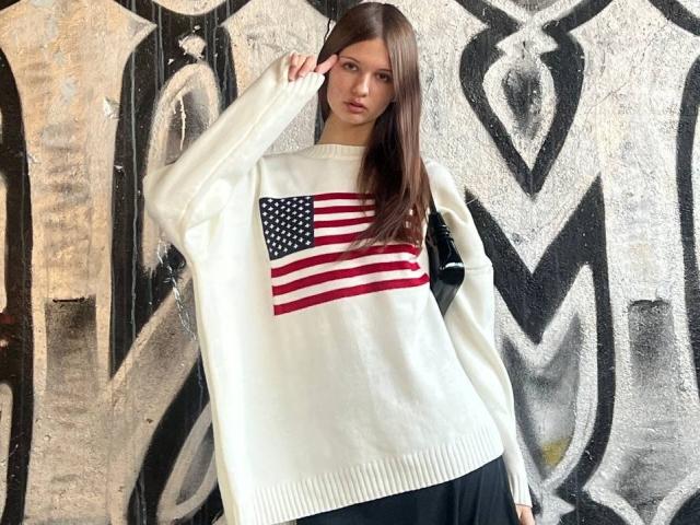 SuzieBrion has long brown hair, wearing a white sweater with a large American flag on the chest, standing in front of graffiti. She's looking at the camera.