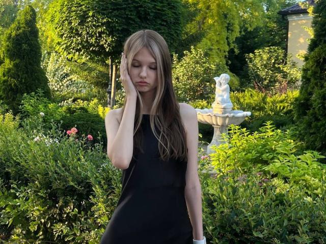 SuzieBrion with long brown hair, wearing a black sleeveless dress, touching her face, standing in a garden with a white statue in the background.