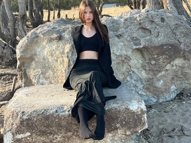 SuzieBrion sitting on rocks, wearing black crop top, black skirt, open jacket, black socks, long brown hair, looking at camera, forest background, daytime, relaxed, sexy.