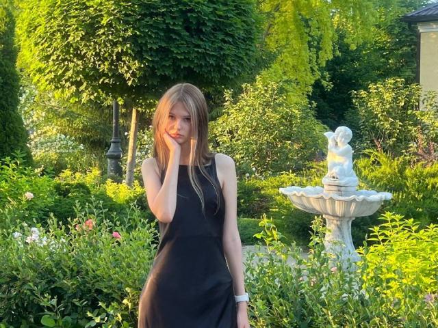 SuzieBrion with long blonde hair, wearing a black dress, stands in a garden, hand near her face, white watch on her wrist, next to a white stone fountain.