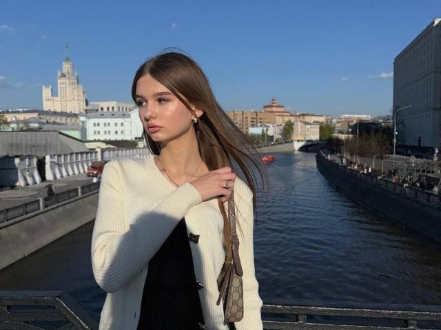 SuzieBrion with long brown hair, wearing a white cardigan and black top, standing by a river, holding a brown bag, looking serious, blue sky, sunlight.
