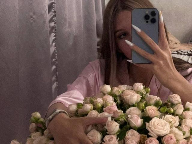 SuzieBrion with pink roses, holding a phone in front of her face, wearing a pink shirt, long blonde hair, white nails, silver bracelet, taking a mirror selfie.