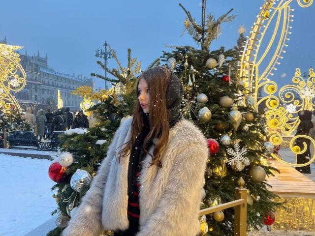 SuzieBrion with long brown hair, wearing a fluffy grey coat and black scarf, stands in front of a decorated Christmas tree with red, silver, and gold ornaments, snowing.