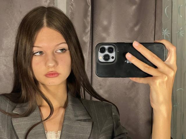 SuzieBrion with long brown hair, gray pinstripe suit, holding a black phone in front of her face, looking slightly bored with pink lips, in a mirror selfie.